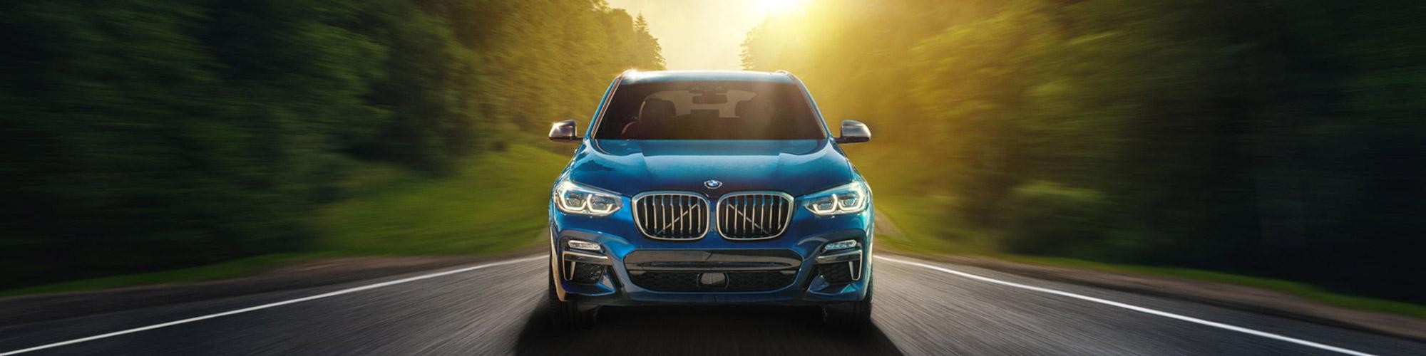 Head-on view of a blue BMW X3 driving on a highway and backlit by the sunset.