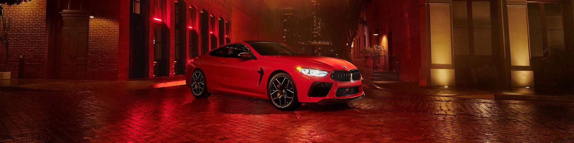 A red BMW M8 parked under a light on a deserted city street.
