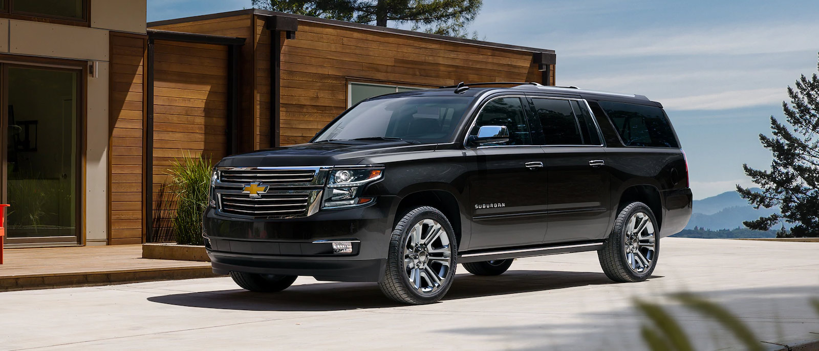 A black 2025 Chevrolet Suburban parked near a scenic shoreline in Amsterdam, NY, under a bright blue sky. 