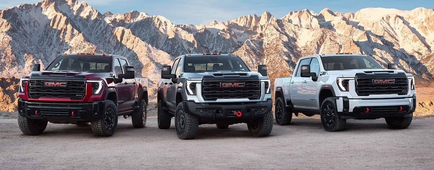 A red, a silver, and a white 2025 GMC Sierra 2500 HD are parked on dirt with a mountain background.
