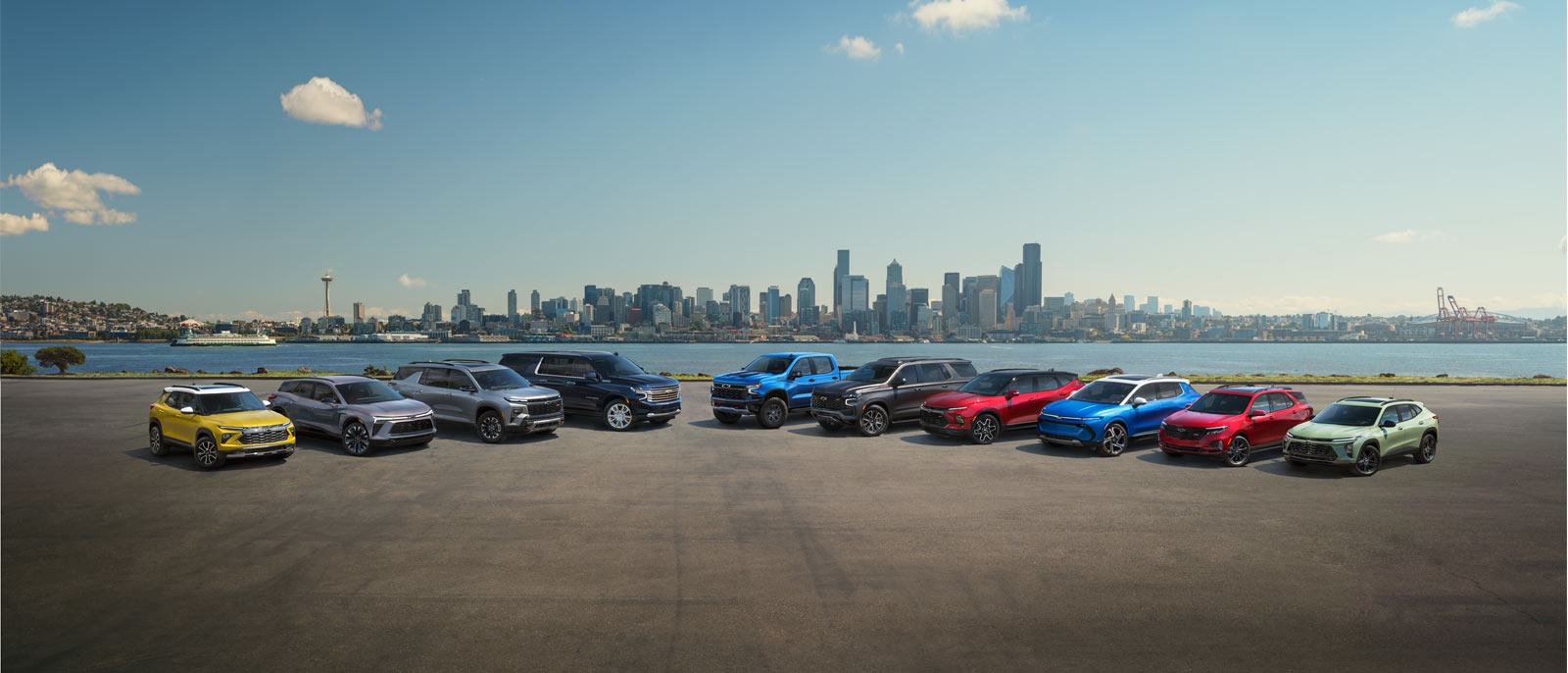 The 2025 Chevrolet lineup parked on a cement port by the water, with downtown Amsterdam, NY in the background near Mangino Chevrolet.