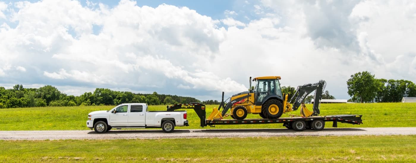 A white 2018 Chevy Silverado 3500HD is towing heavy machinery on a big trailer.