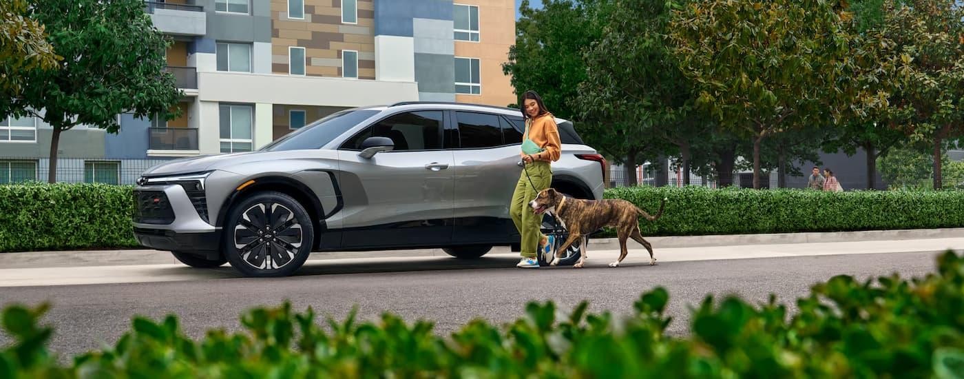 A grey 2025 Chevy Blazer EV RS is parked on the side of a city street as a woman and a dog jog by.