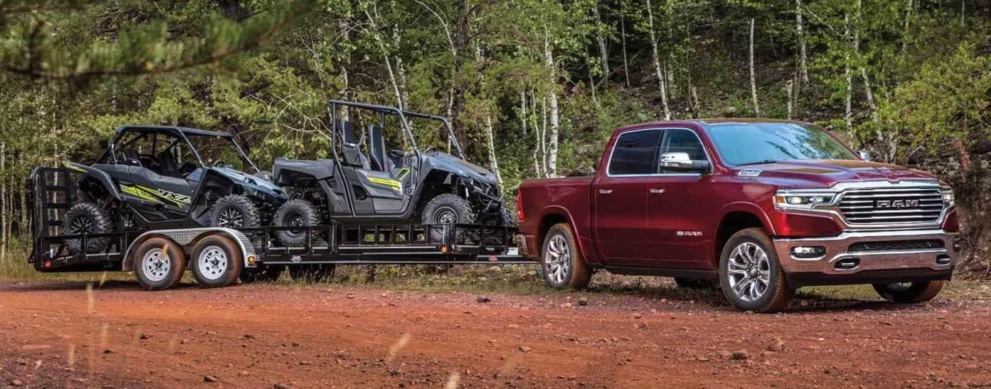 A red 2023 Ram 1500 is towing ATVs on a trailer near a dirt trail.