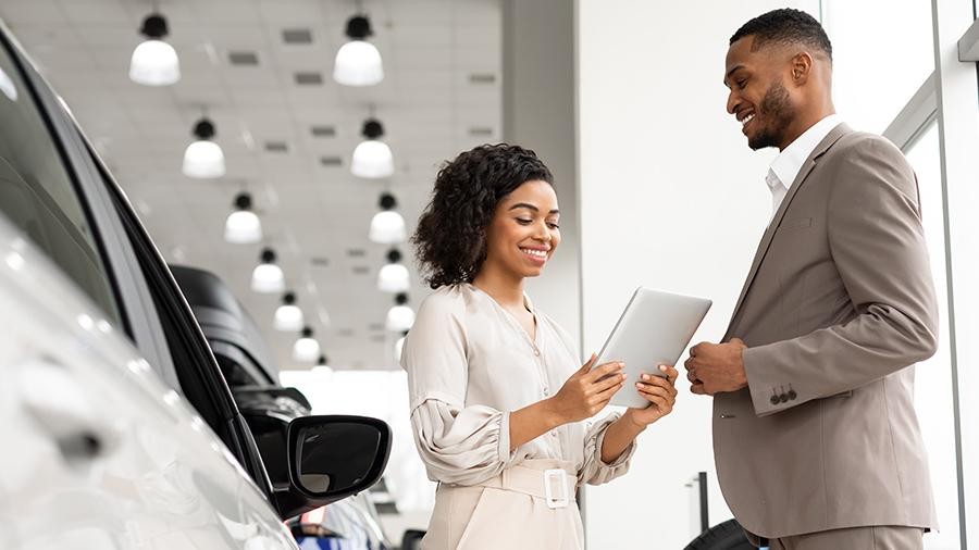 Car Sales. Automobile Seller Woman Showing Auto To African Businessman Standing In Dealership Showroom. Free Space