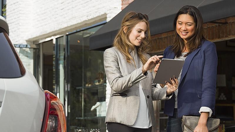 Two women looking at a tablet next to a new car.